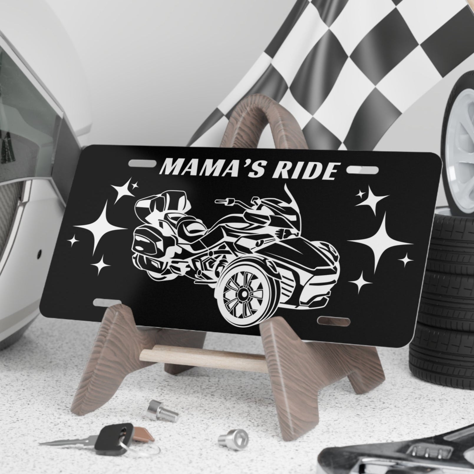 Black license plate with car design and 'Mama's Ride' text, placed on a stand with tools and tires in the background.