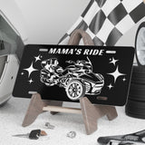 Black license plate with car design and 'Mama's Ride' text, placed on a stand with tools and tires in the background.