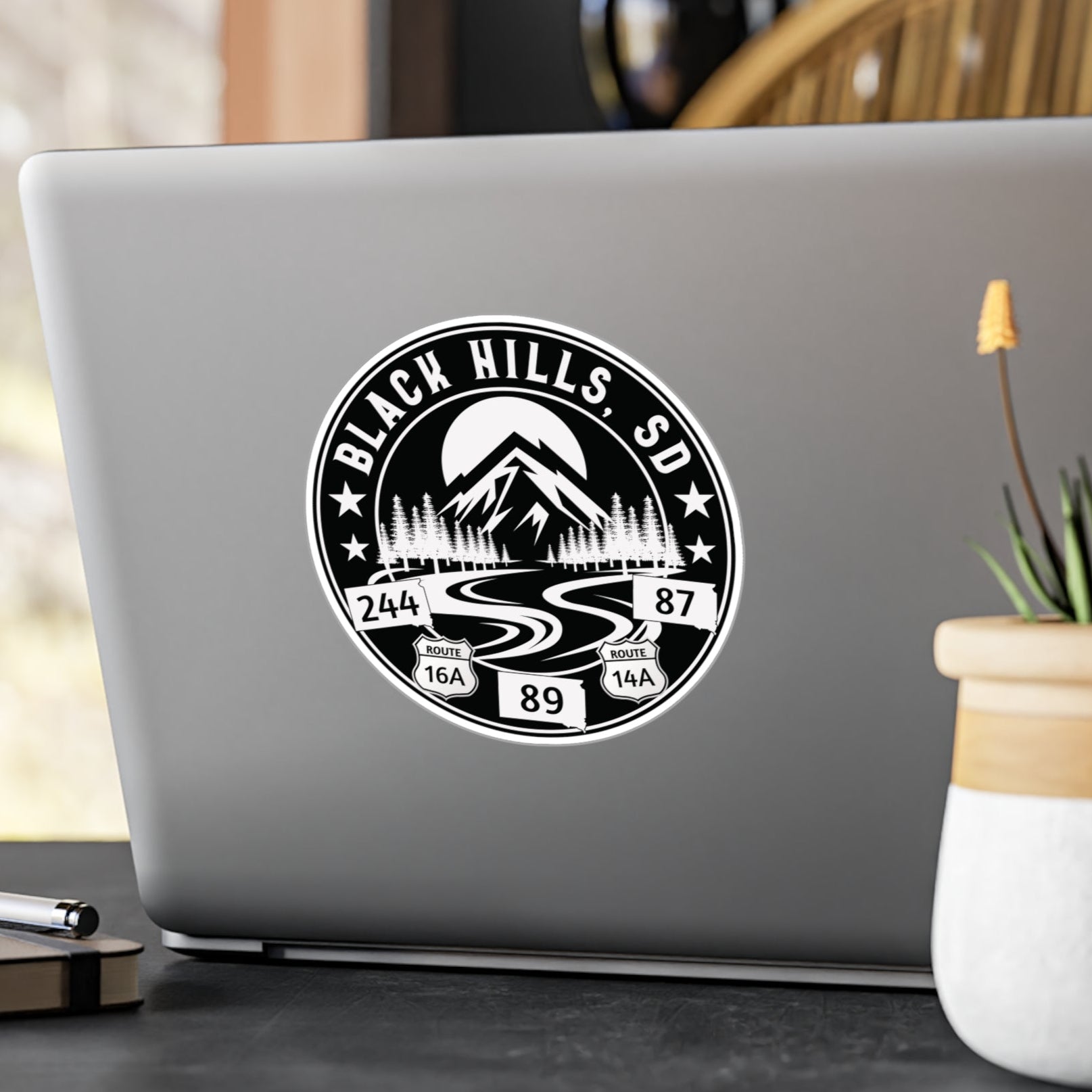 Laptop with a 'Black Hills SD' sticker on a desk with a plant and pen.