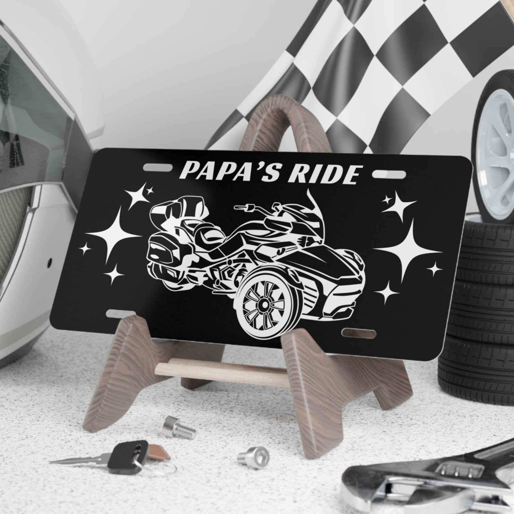 Black license plate with car design and 'Papa's Ride' text, placed on a stand with tools and tires in the background.