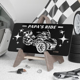 Black license plate with car design and 'Papa's Ride' text, placed on a stand with tools and tires in the background.
