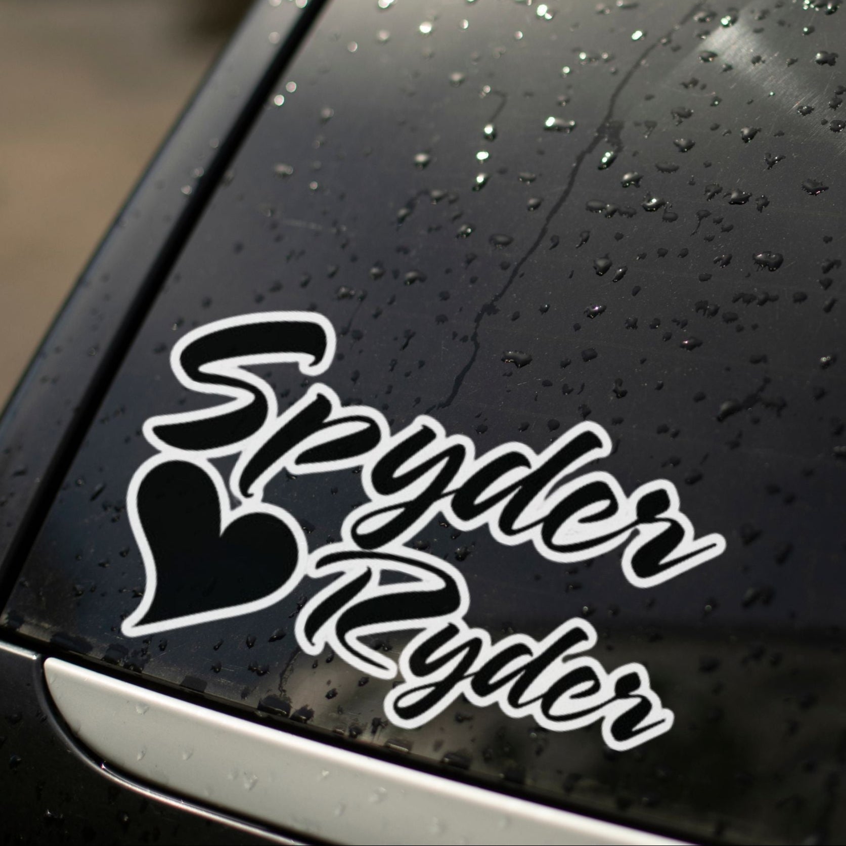 A black decal with the text 'Spyder Ryder' on a car window with rain droplets.