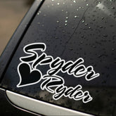 A black decal with the text 'Spyder Ryder' on a car window with rain droplets.