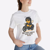 Person wearing a white t-shirt with a bee graphic and text, standing indoors next to a plant.