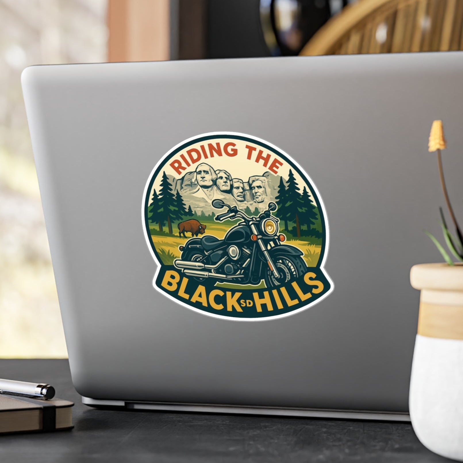 Laptop with a 'Riding the Black Hills' sticker on a desk with a plant and pen.