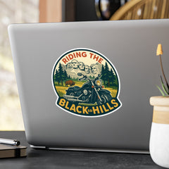 Laptop with a 'Riding the Black Hills' sticker on a desk with a plant and pen.
