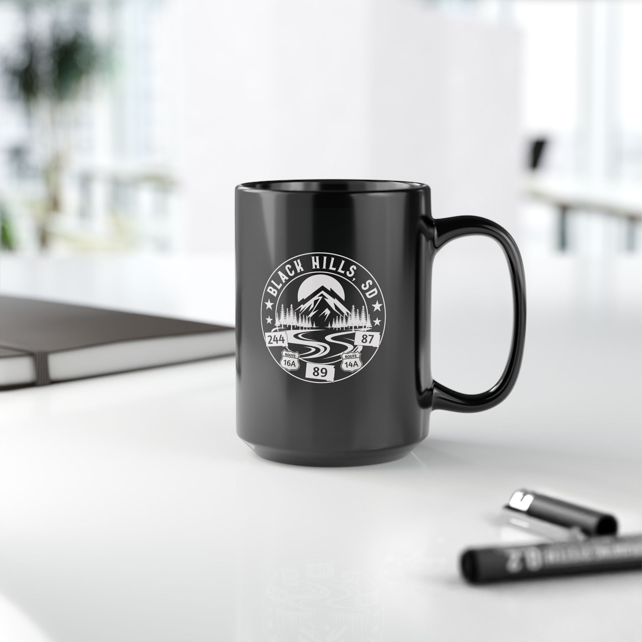 Black mug with 'Black Hills SD' design on a white surface with office items.
