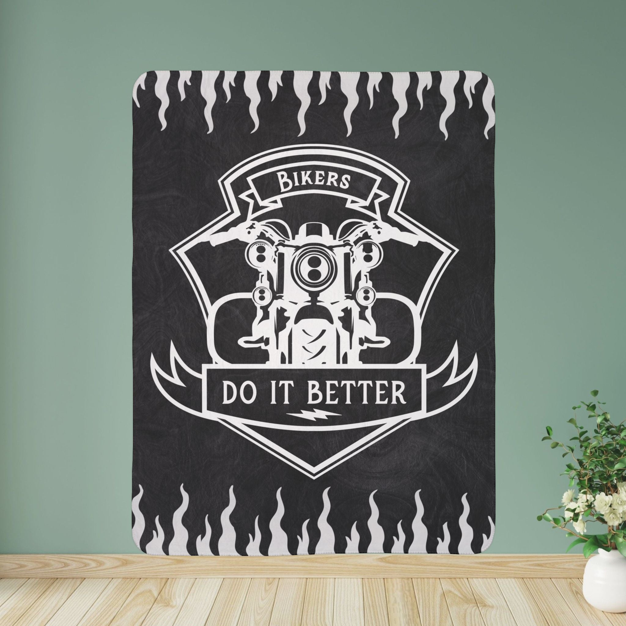 A black Sherpa fleece blanket with a white motorcycle design and the text 'Bikers DO IT BETTER' printed on it, displayed on a wooden surface with a green background.