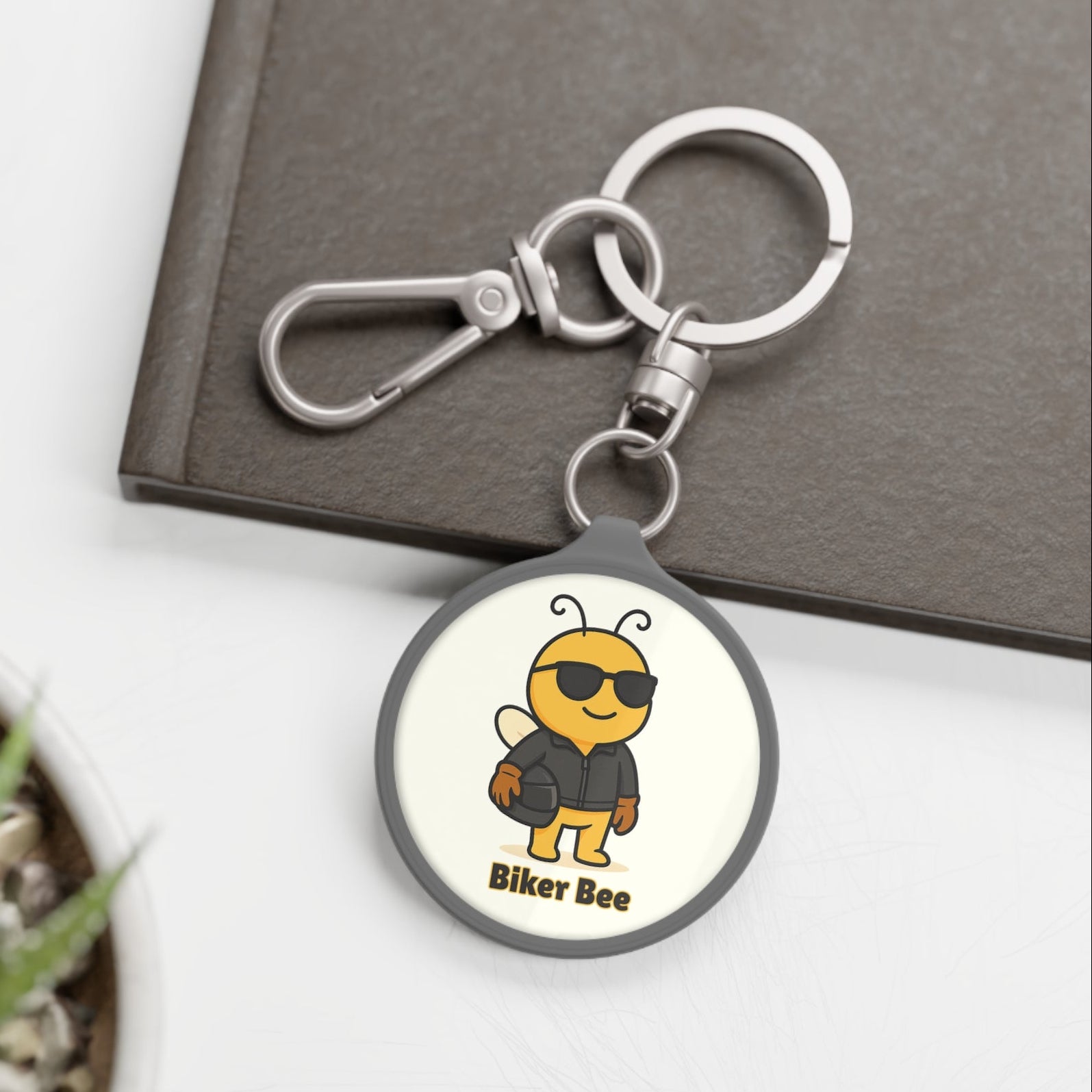 Keychain with a bee design labeled 'Biker Bee' on a white surface with a plant and book in the background.
