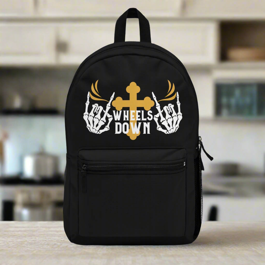 A black polyester backpack with a gold cross and wings design, text 'Wheels Down', and skeleton hand elements on the front pocket.