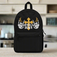 A black polyester backpack with a gold cross and wings design, text 'Wheels Down', and skeleton hand elements on the front pocket.