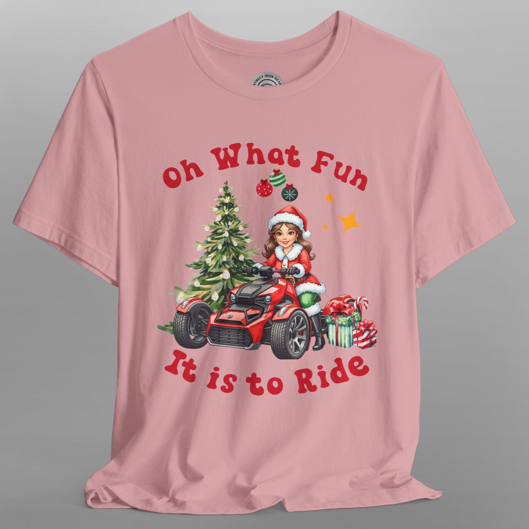 Pink t-shirt with Christmas-themed design featuring a child on a bike with trees and presents, on a gray background.