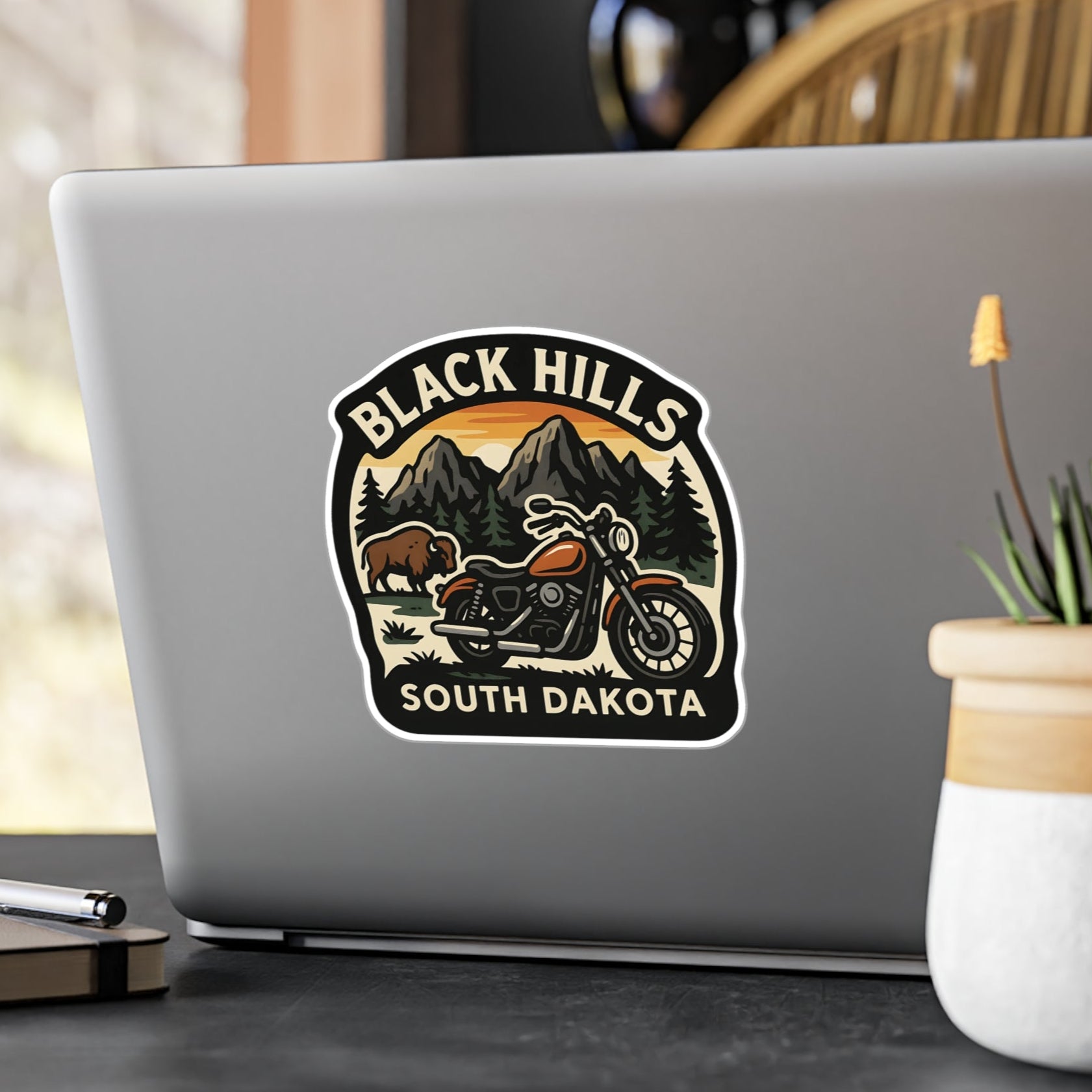 Laptop with a 'Black Hills South Dakota' sticker on a desk with a plant and pen.