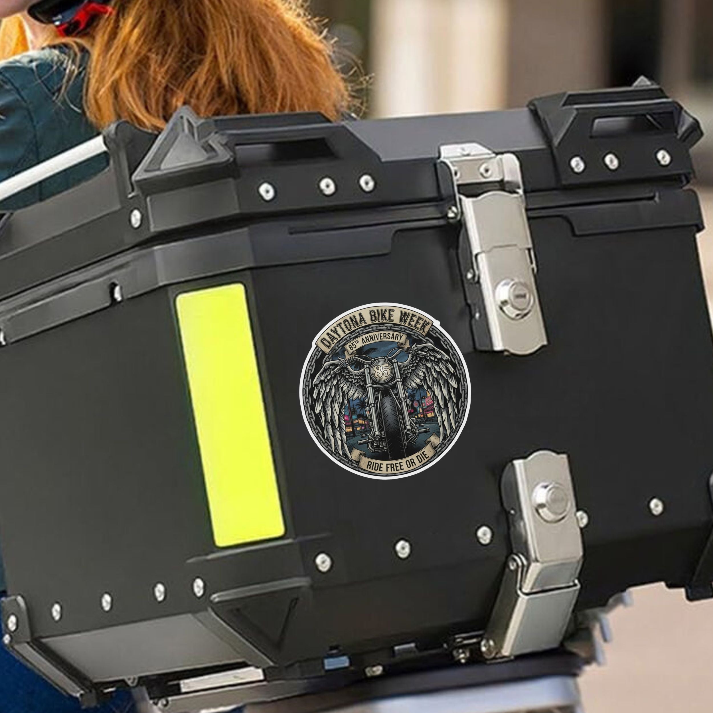 Black motorcycle storage box with a decorative emblem featuring a motorcycle and text, on a blurred background.