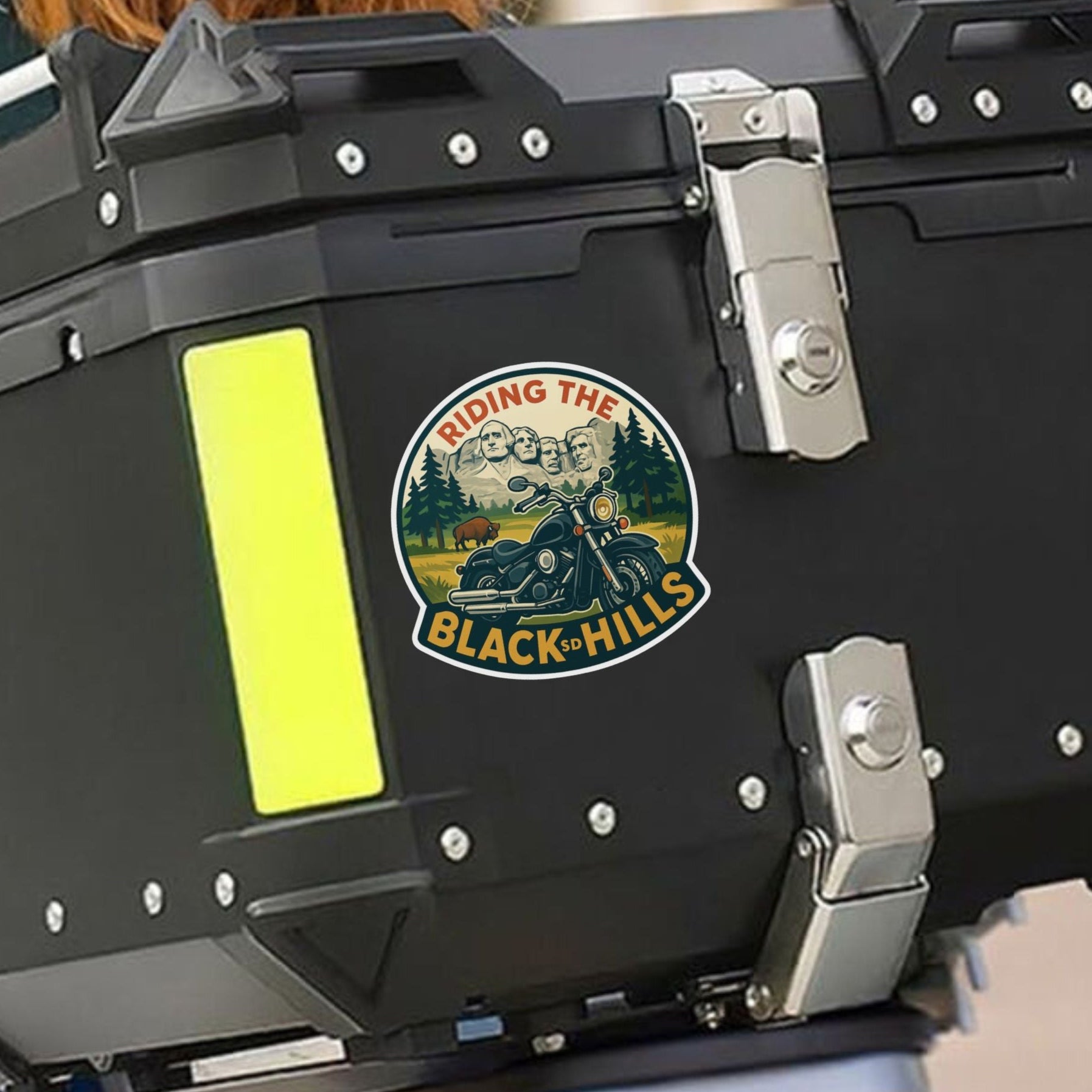 Black motorcycle storage box with 'Riding the Black Hills' logo on a blurred background