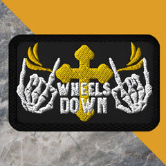 An embroidered patch featuring a design with skeleton hands, a gold cross with wings, and the text 'WHEELS DOWN'. The patch is black with yellow and white accents.