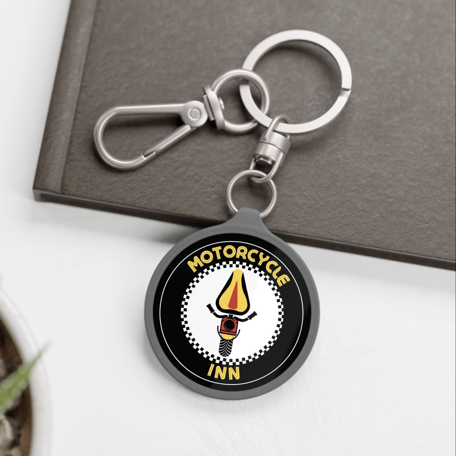 Keychain with 'Motorcycle Inn' logo on a white surface with a plant and book in the background